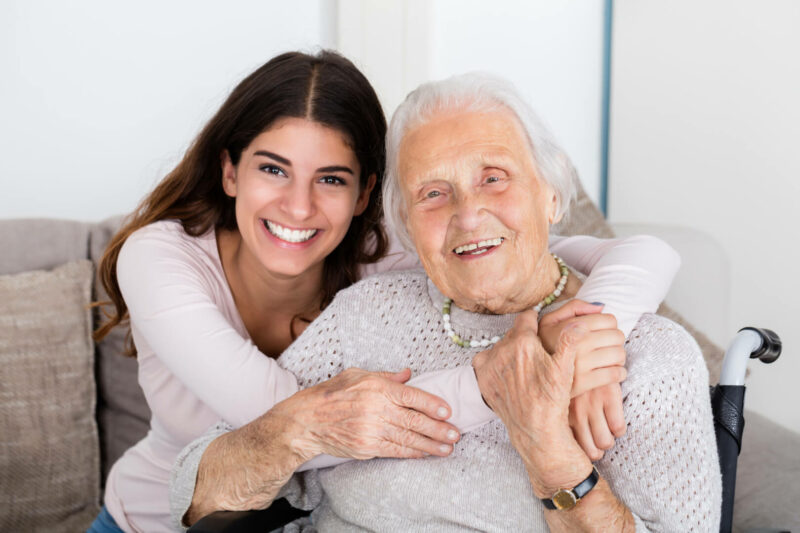 En la imagen aparece una cuidadora joven abrazando a una señora mayor con mucho cariño desde atrás, ambas sentadas en un sofá y posando para la cámara.