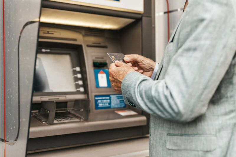 Un señor mayor mira su tarjeta de crédito para sacar dinero en un cajero automático. No o se aclara con la tarjeta y necesita la ayuda de una persona del banco que le atienda. .