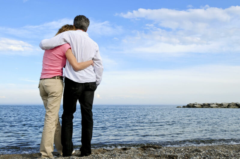 En la imagen aparece un matrimonio de personas adultas, de la generación baby boomer, en una playa, abrazados de espaldas mientras miran el agua.