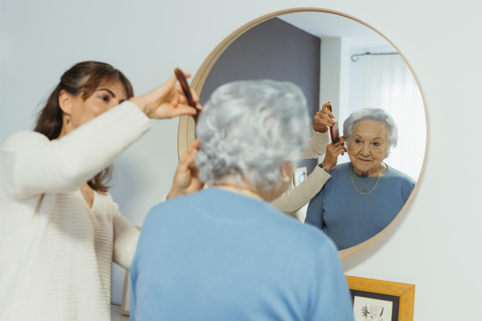 Cuidadora ayudando a que una señora mayor se peine mientras esta se ve en un espejo.