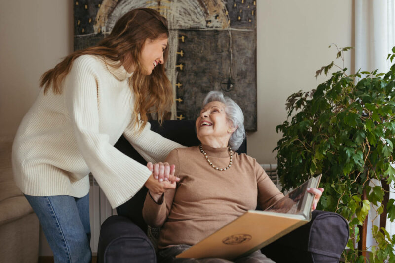 Una cuidadora muestra cariño a una anciana que se encuentra sentada mirando un álbum de fotos en el sofá de su casa. 