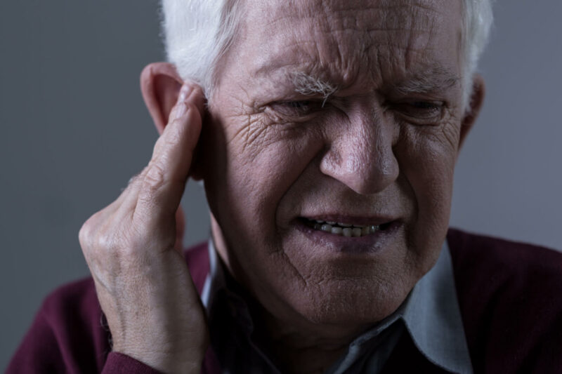 Un anciano se queja de un dolor de oído mientras posa su mano en el mismo y expresa dolor facialmente. 