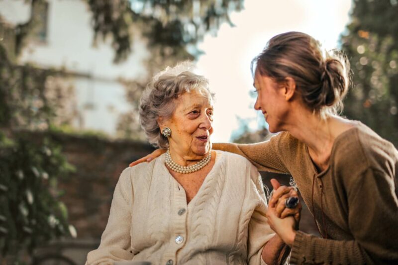 En la imagen aparece una persona mayor, una mujer, charlando con su hija, que parece ser su cuidadora doméstica, en el exterior, en una terraza de una casa con jardín. 