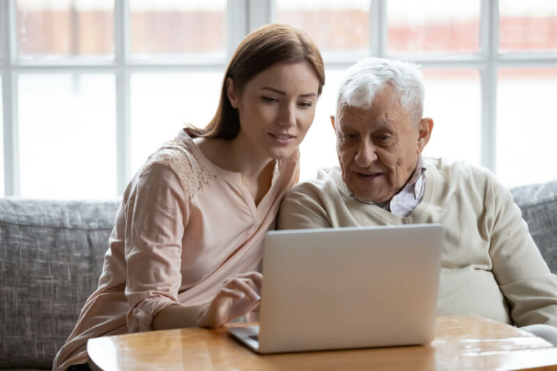 En la imagen aparece una cuidadora ayudando a manejar el ordenador a un hombre mayor en el sofá de una casa.