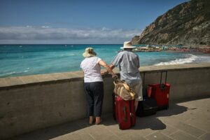 Dos personas mayores de espalda observando el mar desde una acera con sus maletas