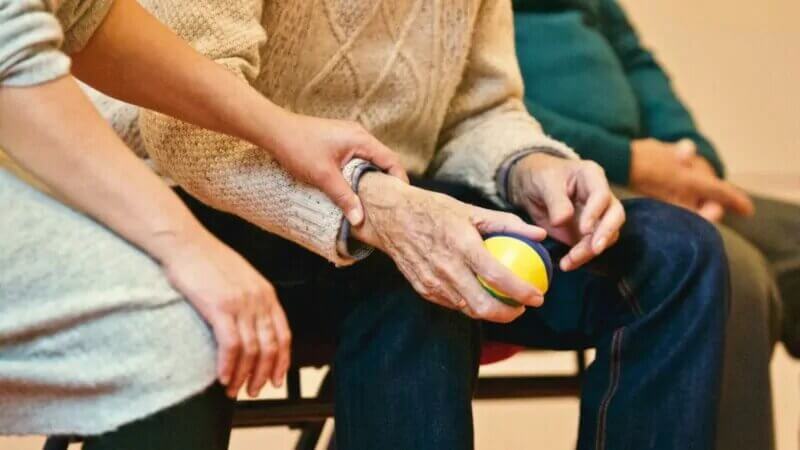 Una cuidadora interna agarra de la mano a una persona mayor mientras sostiene una pelota de goma. Es un primer plano de ambas manos.