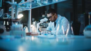 Hombre joven con gafas y bata blanca, en un laboratorio, mirando una muestra por un microscopio.