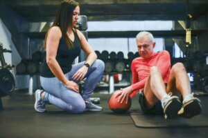 Un hombre mayor en el gimnasio con su entrenadora personal. Ella está de rodillas viendo como el hombre, tumbado en la esterilla, hace ejercicios con un balón medicinal de color rojo y negro.