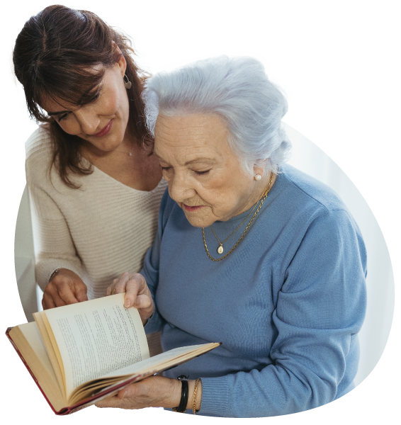 Señora mayor sosteniendo un libro abierto mientras a su lado derecho permanece su cuidadora.