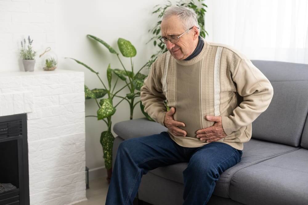 Un señor mayor sentado en el sofá de su casa ante un dolor estomacal. 