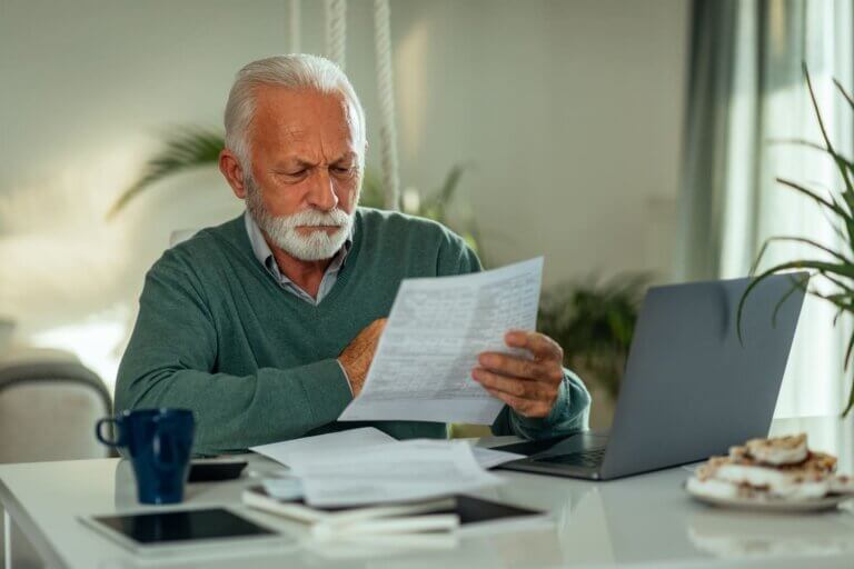 Un hombre mayor analiza las facturas de su banco desde su casa con un portátil.