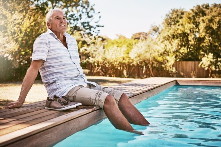 Un hombre mayor entado en el bordillo de una piscina al sol.