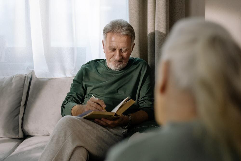 Un hombre mayor sentado en su sofá escribe algo en su cuaderno. 