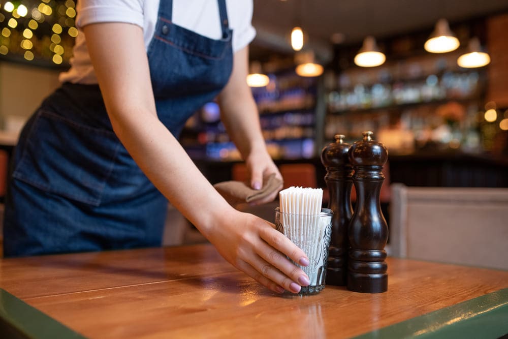 Una camarera con delantal deposita un vaso con palillos de madera en una mesa.