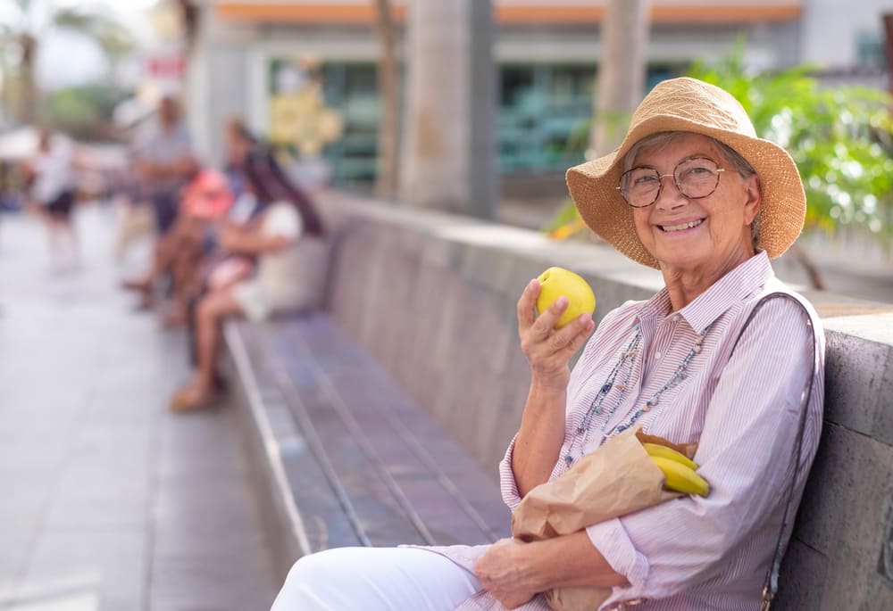 Una mujer mayor está sentada en un banco a la sombra mientras come una manzana. 