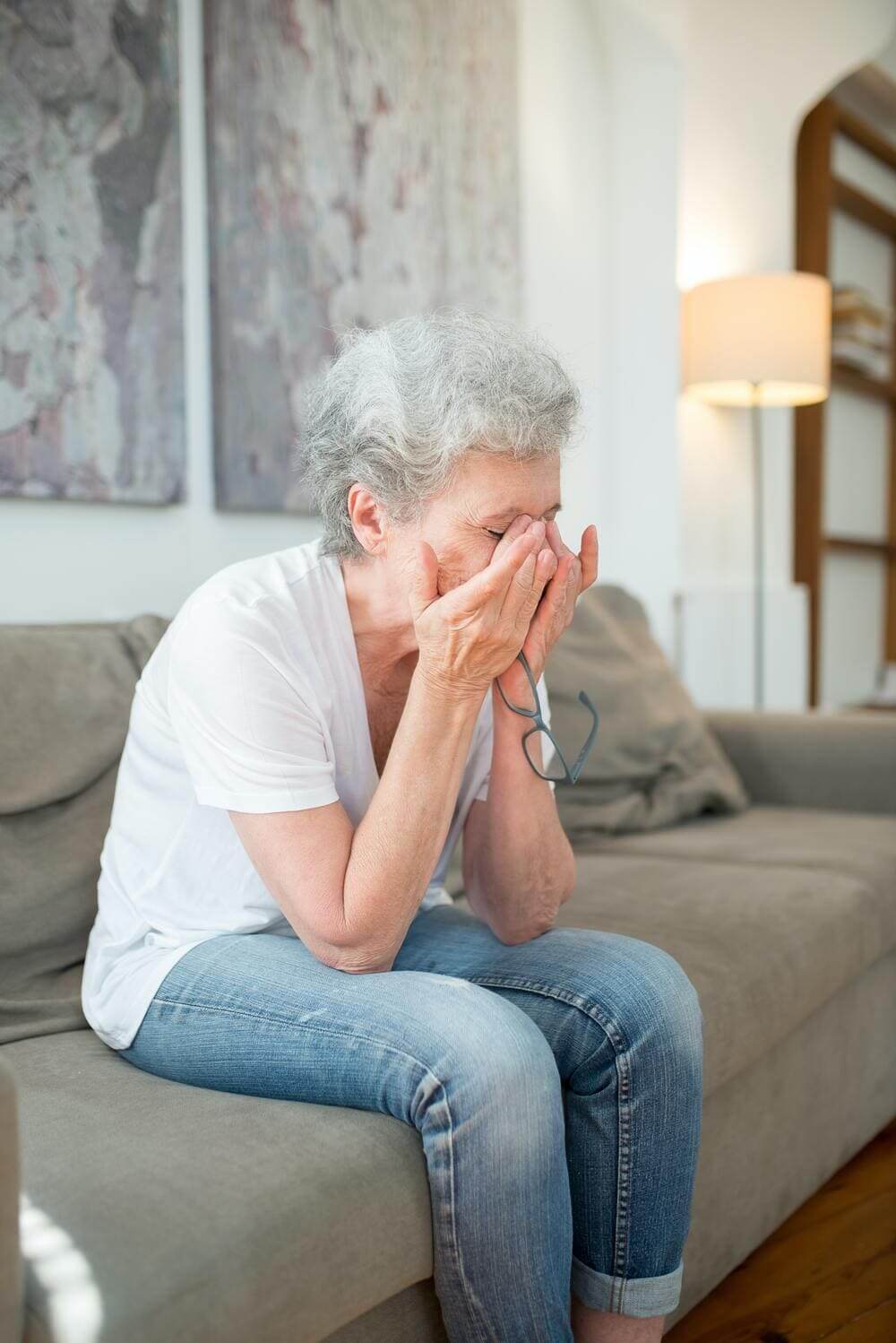 Una imagen de una mujer mayor sentada en el sofá de su casa rascándose los ojos ante problemas de visión. 