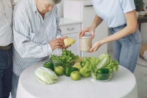 Una persona mayor con su cuidadora sosteniendo verduras de una mesa.