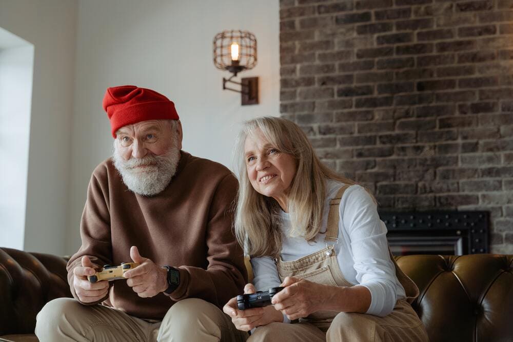 Un abuelo y una abuela están jugando videojuegos sentados en el sofá de su casa.