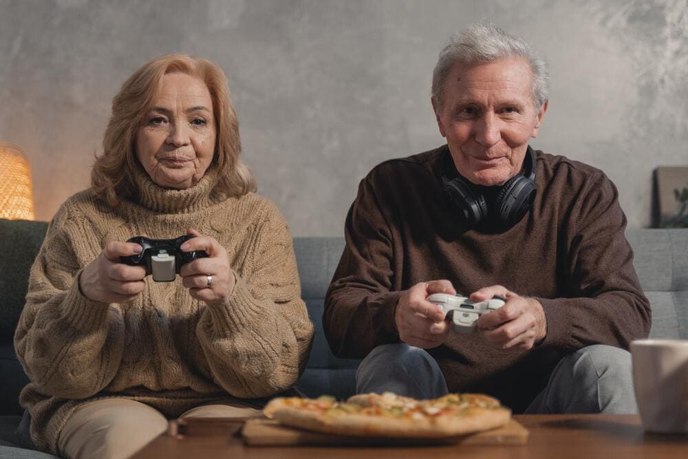 Un abuelo y una abuela juegan a videojuegos sentados en su sofá y en la mesa tienen una pizza.