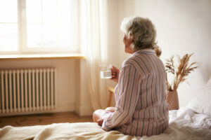 Una anciana sentada en el borde de la cama con un vaso de agua en la mano