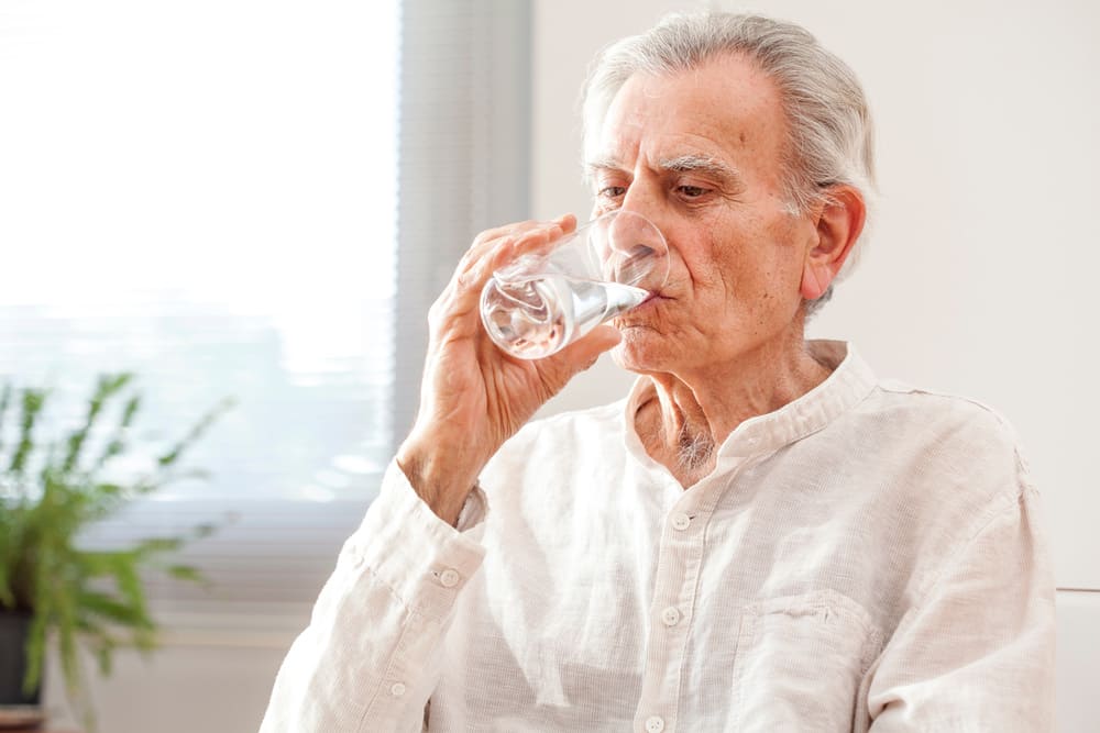 Un anciano bebe agua de un vaso en su casa. 