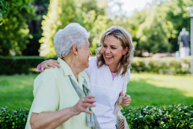Una joven pasea por el parque con una señora mayor que cuida mientras que muestran sonrientes.