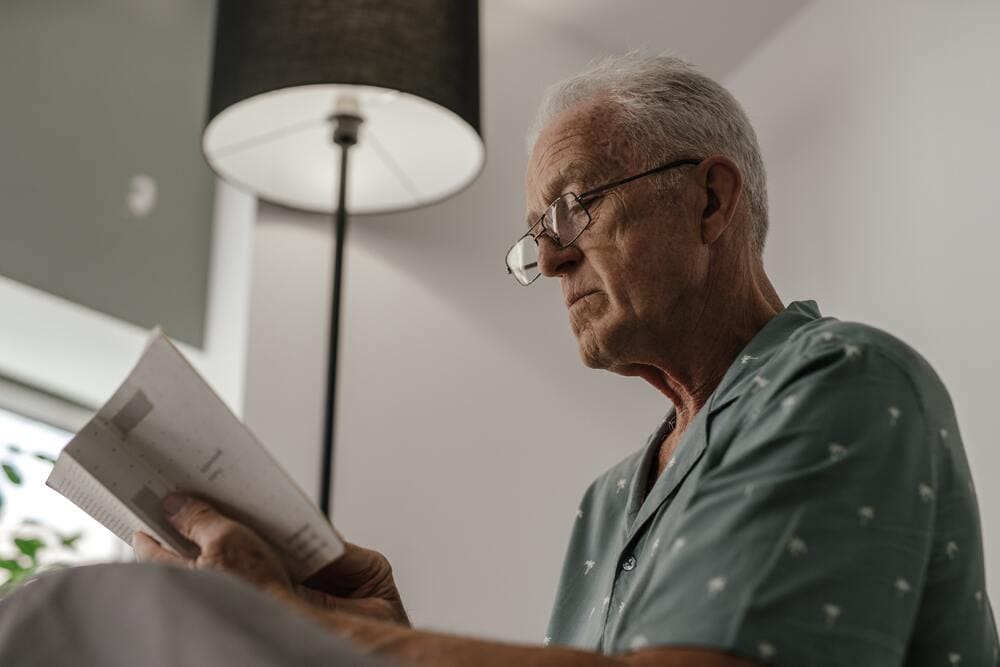 Un señor mayor lee un libro en el salón de su casa. 