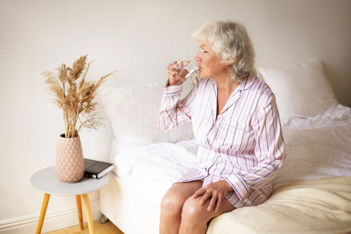 Una anciana en la cama bebiendo un vaso de agua.