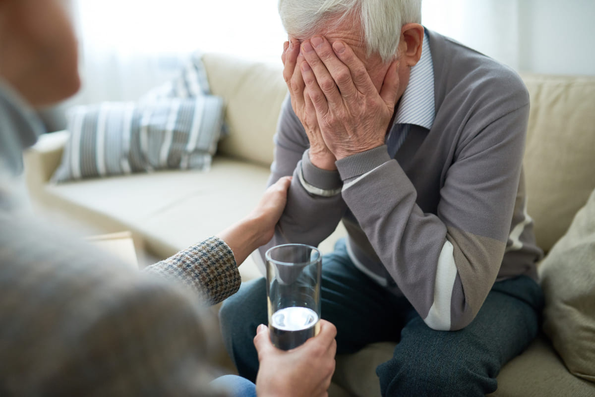 Un anciano llorando sentado en un sofá, mientras alguien lo consuela y le da un vaso de agua.