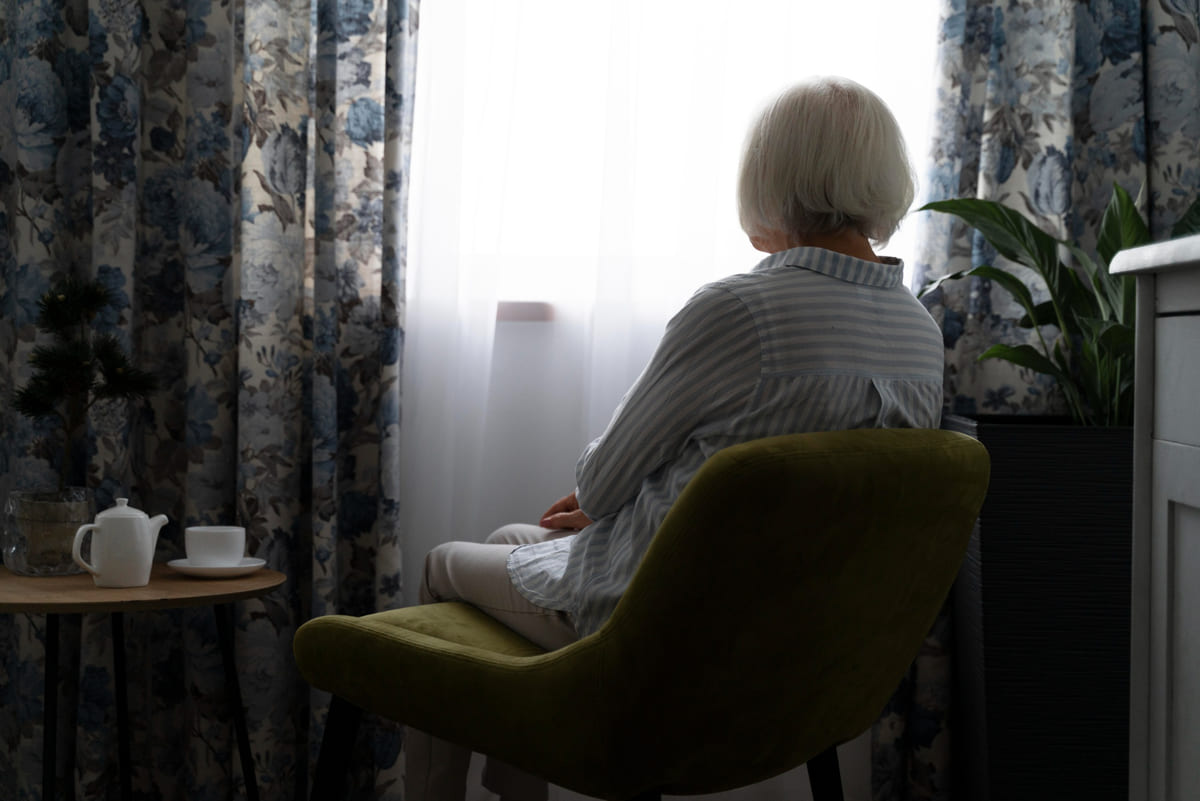 Una anciana sola sentada en una silla frente a una ventana.