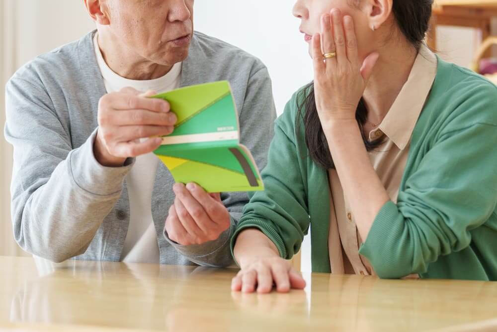 Una pareja de jubilados discute en una mesa mientras el hombre le enseña la cartilla del banco a la mujer. 