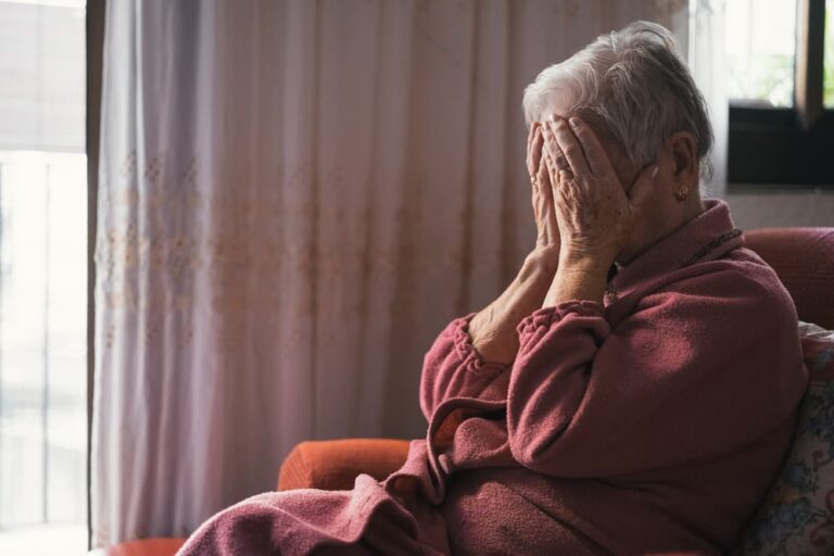 Una mujer de edad avanzada se tapa la cara ante un episodio de neuralgia del trigémino en el sofá de su casa.