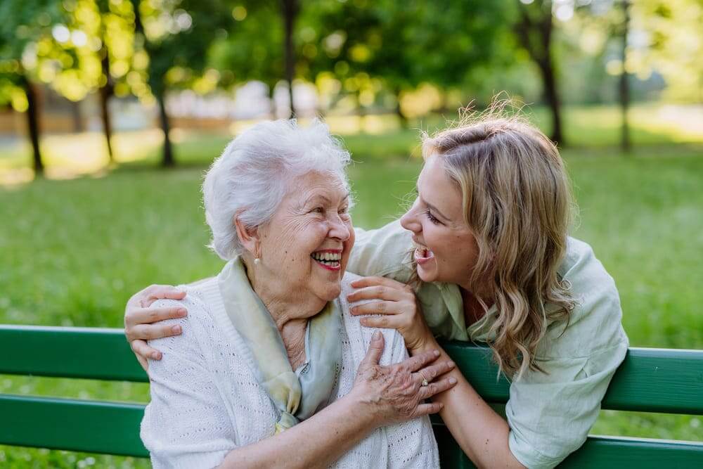Una chica joven sonríe mientras saluda a una anciana en un banco de un parque. 