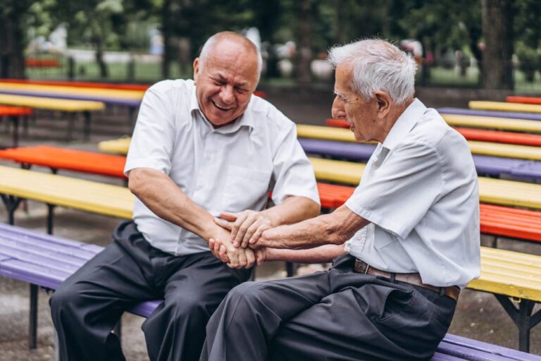 Dos hombres mayores saludándose y riéndose en un banco.