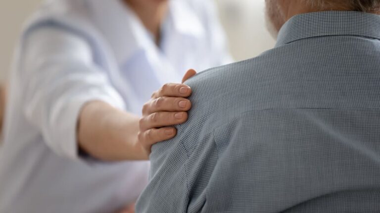 Una cuidadora apoya su brazo en claro gesto de saludo en el hombro de un anciano.