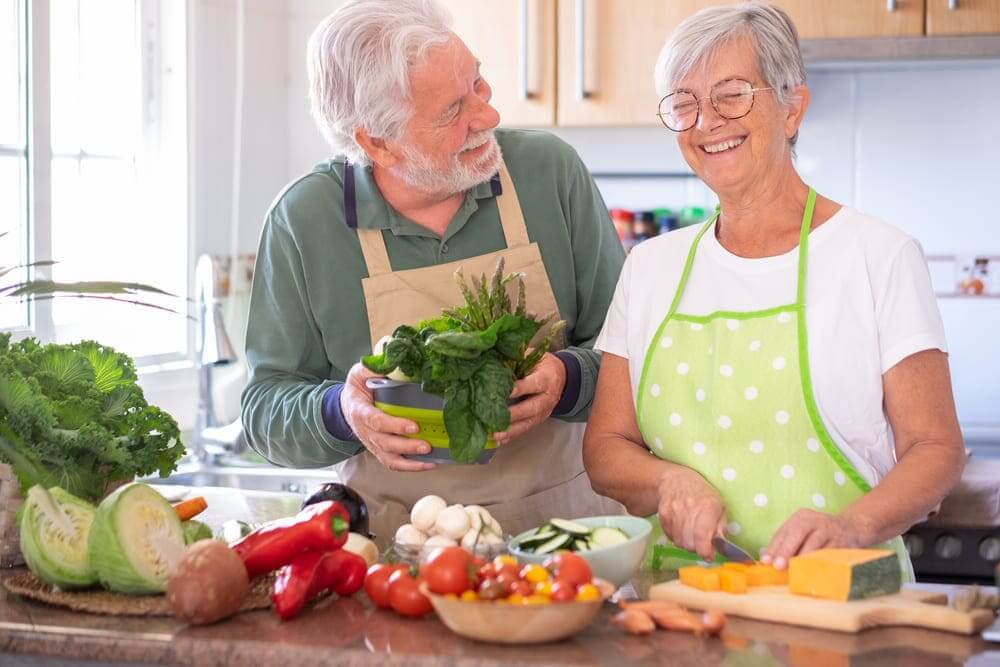 Dos personas mayores cocinan con verduras mientas sonríen en la cocina. 