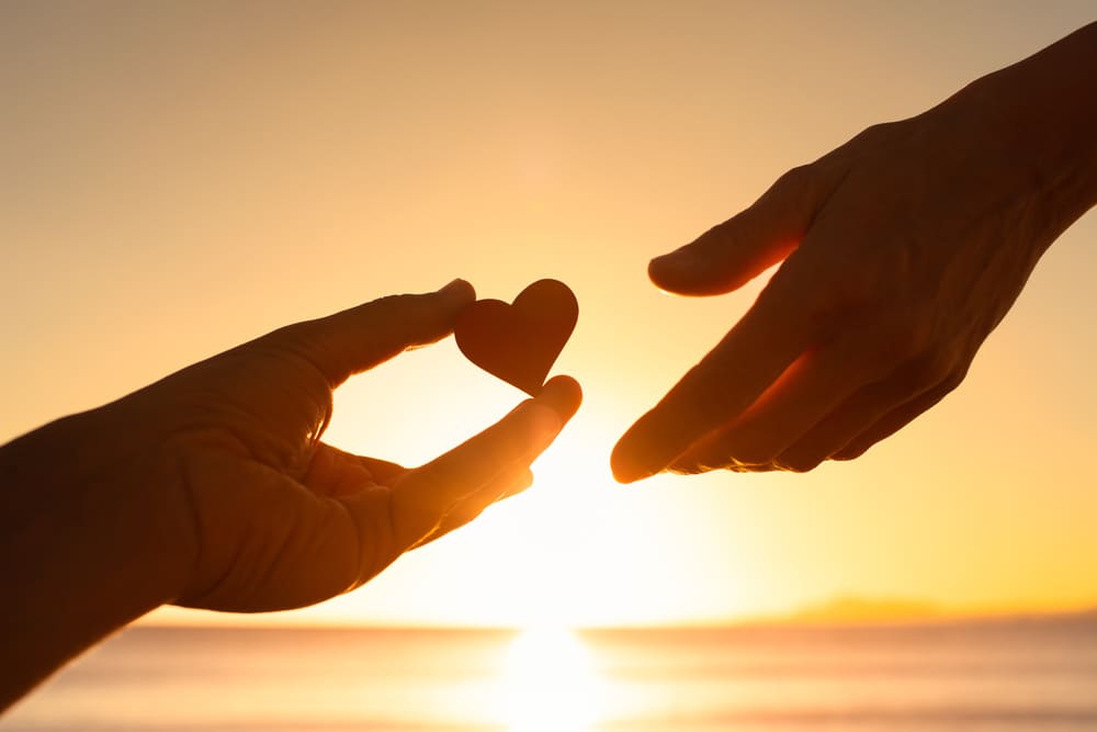 Una imagen de dos manos en la playa, una de ellas le entrega un corazón de papel a otra mano. 