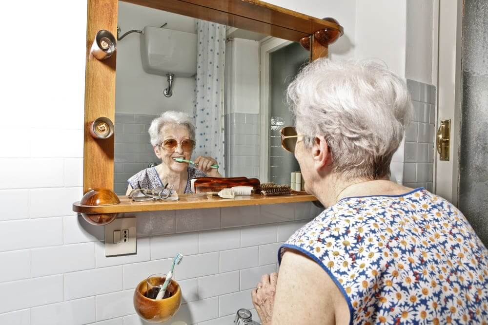 Una anciana se lava los dientes mientras se mira en el espejo de su lavabo. 