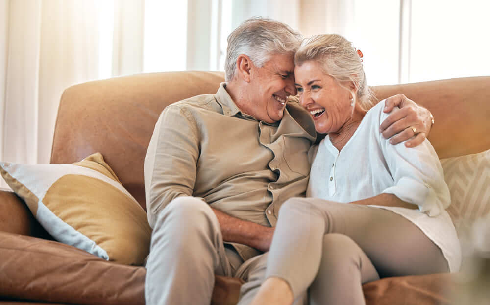 Una pareja de ancianos sentados en su sofá ríen entre ellos. 