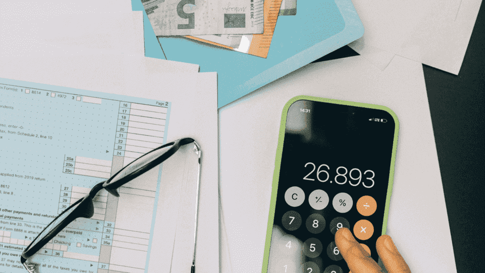 Un móvil con la calculadora junto a unas gafas en una mesa y unos billetes de euro. 
