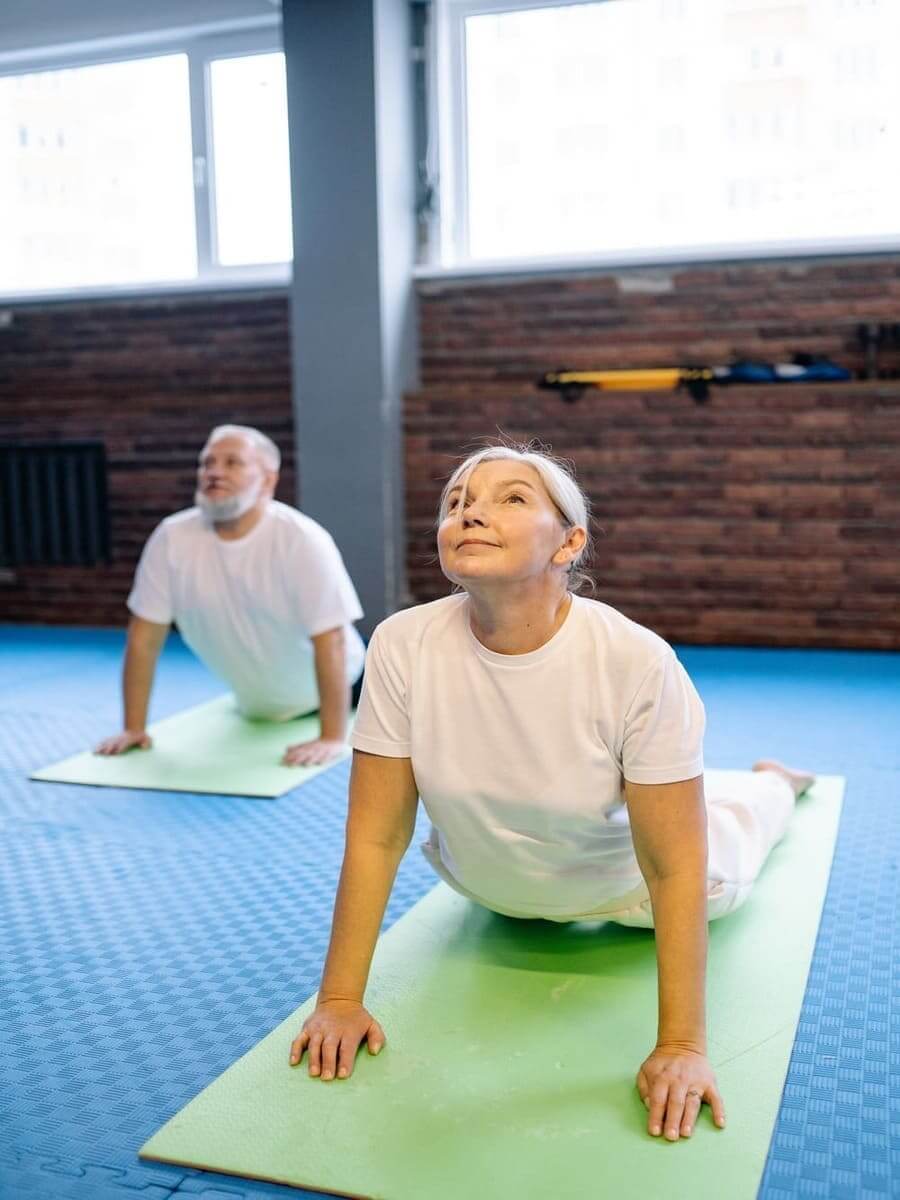 Dos personas mayores en un gimnasio con una esterilla haciendo estiramientos. 