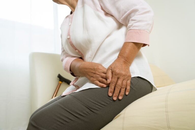 Una señora se queja de un dolor de cadera sentada en su cama.
