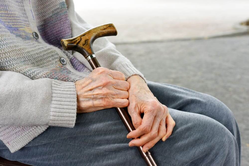 Un plano tres cuartos de una persona mayor sentada en un banco con su bastón. 