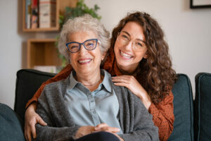 Mujer de edad avanzada sentada en un sofá sonriendo. Por detrás se asoma su nieta, abrazándola.