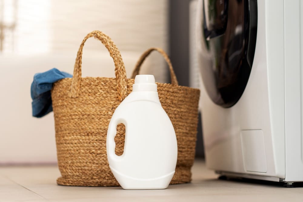 Una cesta de mimbre con ropa junto con un bote de detergente y una lavadora. 