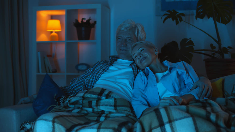 Dos ancianos de noche viendo la teletienda en la televisión en el sofá de su casa. 