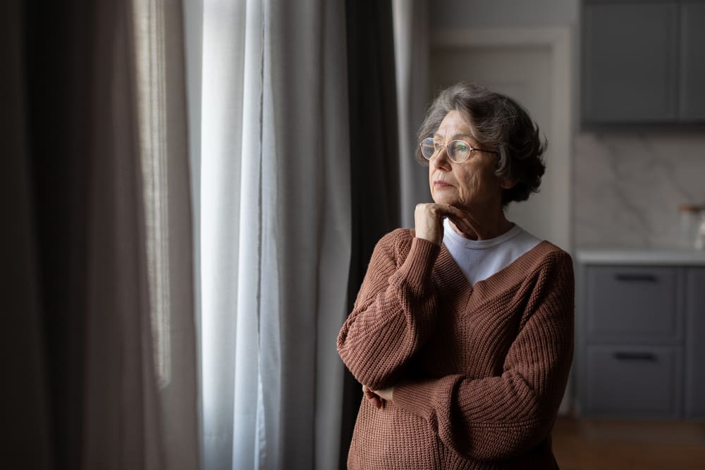 Una anciana en soledad mira con la mirada perdida la ventana de su casa que da al exterior. 