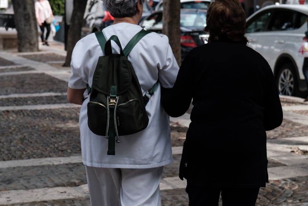 Una cuidadora coge del brazo a una anciana mientras pasean por la calle.