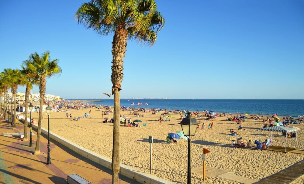 Una panorámica de la playa de La Costilla en Cádiz, con mucha gente disfrutando en la arena y en un día soleado. 