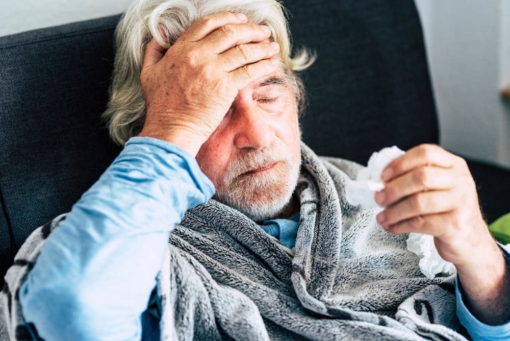 Un señor mayor sentado en el sofá se toca la cabeza ante la fiebre. 